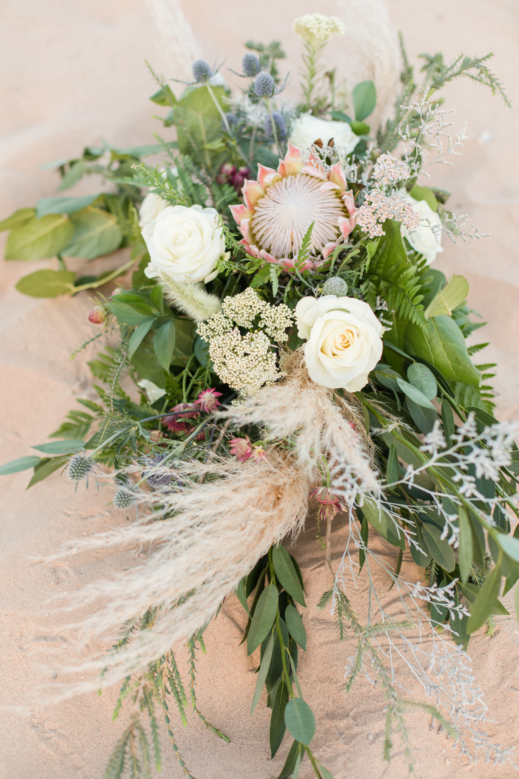 A protea and white rose bouquet resting on sand